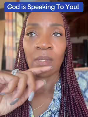 A close-up shot of a woman with long, braided, burgundy hair. She has a serious expression, and her finger is touching her lips suggesting she is about to speak or is making a point.