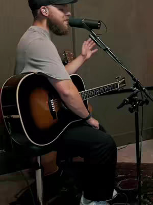 A man with a beard, wearing a grey t-shirt, black cap, and black pants, is shown seated while playing an acoustic guitar. He is in a room with minimal decor and a microphone in front of him.
