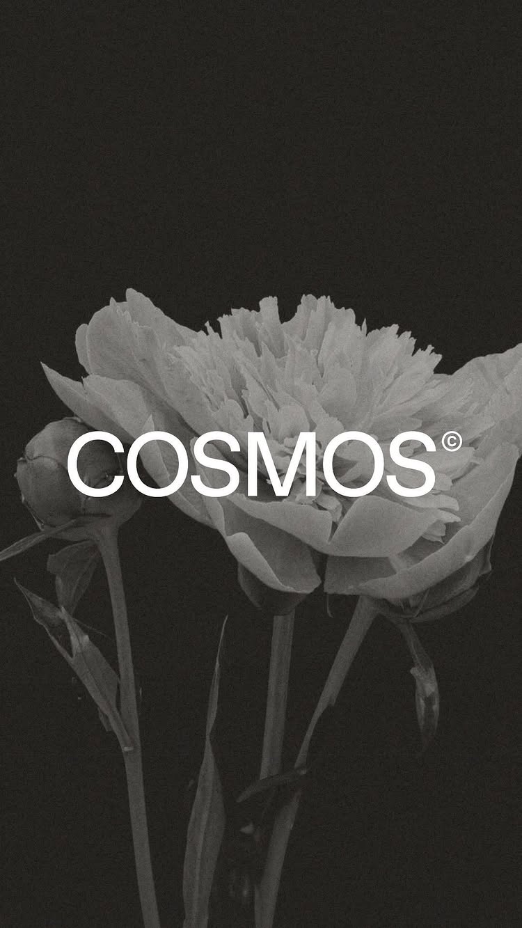 A stark black and white image featuring a close-up of a delicate white flower, likely a peony, set against a completely black background. The flower is the central focus, with its soft, layered petals prominently displayed. The word "COSMOS" is overlaid on the flower in a bold, white sans-serif font, with a small registered trademark symbol at the end. The image's simplicity and high contrast create a clean, modern aesthetic.