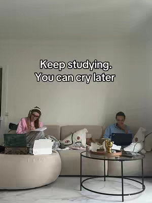 The image shows two people studying in a well-lit living room. One person sits on a large round ottoman on the left side of the image, writing in a notebook. The other person sits on a neutral-colored couch on the right, working on a laptop. There's a coffee table in between and the text overlay says 'Keep studying. You can cry later'.
