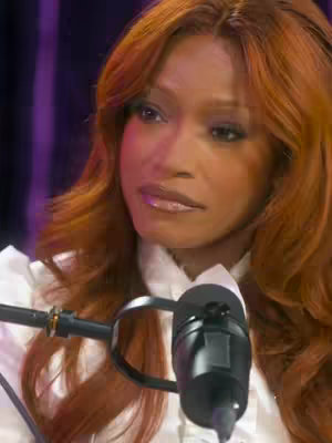 A medium shot captures Keke Palmer with auburn hair styled away from her face, looking slightly off-camera to her left. She wears a white top with ruffles and is seated, with a microphone positioned in front of her. The background is a blurred mix of warm tones, suggesting a studio setting. The lighting is soft, emphasizing Palmer's facial features.