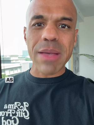 A close-up shot of a man with a shaved head, the focus on his face as he looks directly at the camera. He's wearing a black t-shirt with some words printed in white script. Some building are visible in the background.