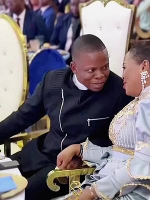 A medium shot shows a man and a woman sitting at a table. They both appear to be in an intimate conversation, with the man speaking to the woman with a warm expression while touching her hand. The shot is taken from the camera's perspective, at eye level, with the focus primarily on the couple. The background shows a blurred, ornate interior with gilded details that suggest a formal setting.