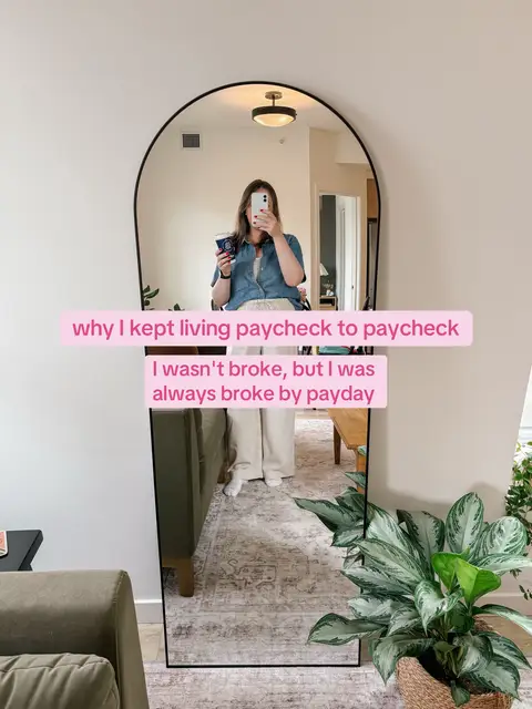A woman is reflected in a large arched mirror, holding a cup. Text overlays on the mirror read, 'why I kept living paycheck to paycheck,' with subtext about being always broke. The setting appears to be a brightly lit interior with modern decor and a green sofa.