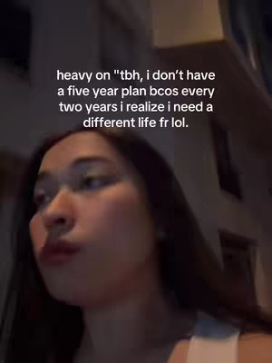 A selfie-style shot of a young woman from the chest up, looking slightly off-camera. Text overlayed on the image states a personal sentiment about not having a five-year plan. The background is blurred suggesting an indoor setting, the colors are muted and overall the image has a casual feel.