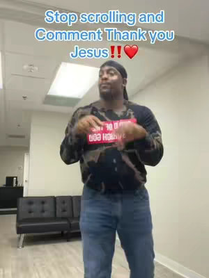 The cover image features a Black man standing in an office setting. He is wearing a black and white patterned long-sleeve shirt, blue jeans, and a black bandana. The text overlay is in a bold, sans-serif font in light blue and white, asking viewers to comment "Thank you Jesus" with a red heart emoji. The background consists of a white wall, with an apparent ceiling lighting fixture. The man is positioned centrally, making a gesture with his hands.