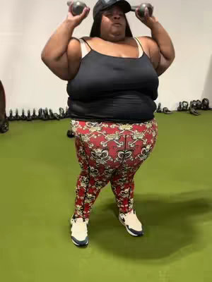 A full-body shot of a woman in a gym setting, wearing a black tank top, patterned leggings, and a baseball cap. She's holding two dumbbells overhead. The background is a plain green surface with exercise equipment visible.