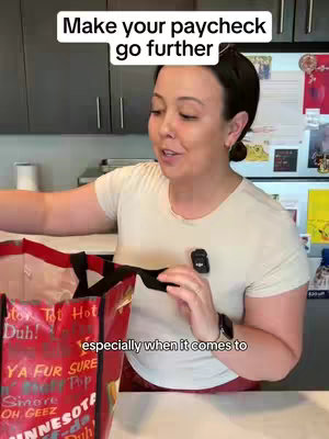 A woman in a white t-shirt is shown from the mid-chest up, near a kitchen counter, reaching for a shopping bag. Text reading "Make your paycheck go further" is displayed at the top of the video. The image is well-lit with a modern design.