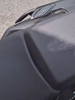The image shows a partial view of a motorcycle seat with a grab handle. The seat appears to be made of dark gray, textured material, likely leather or a synthetic alternative. The grab handle is a sleek, metallic gray, positioned at the rear of the seat. The angle of the shot emphasizes the texture and shape of the seat, and the neutral tones suggest a focus on the vehicle's design.