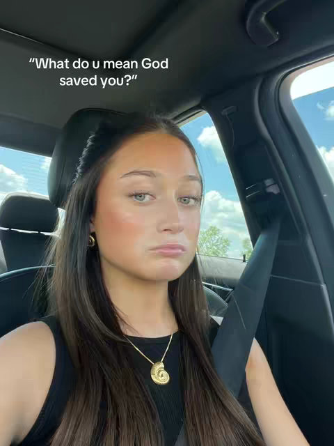 The cover image is a selfie taken from inside a car. A young woman with shoulder length brown hair is centered in the frame, looking directly at the camera with a neutral expression. She wears a black top and a gold necklace. Text overlaid on the image asks, "What do u mean God saved you?"
