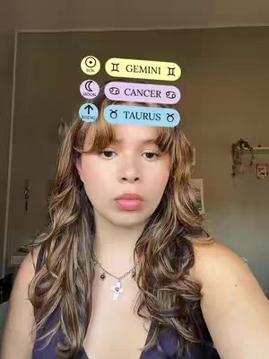 A medium shot featuring a young woman with long, wavy hair, wearing a black top and a necklace. Above her head are several rounded rectangular overlays with text and symbols. The background is a neutral wall. The overall composition is centered and focused on the woman's face.