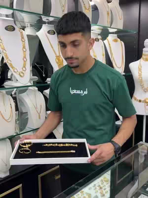 A man in a green t-shirt holding a tray with jewelry, set in front of glass shelves filled with more jewelry pieces. The man has short dark hair and a short beard. The background is filled with various necklaces and bracelets displayed on white stands.