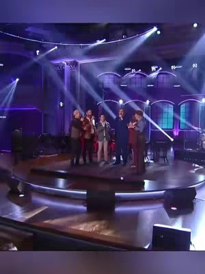 A group of men are standing on a stage under bright lights. The stage has a circular shape with a dark brown wooden texture. Soft, purple lighting emanates from the background, where architectural designs are visible. The men on stage are dressed in various outfits. Most are wearing suits or sport coats.