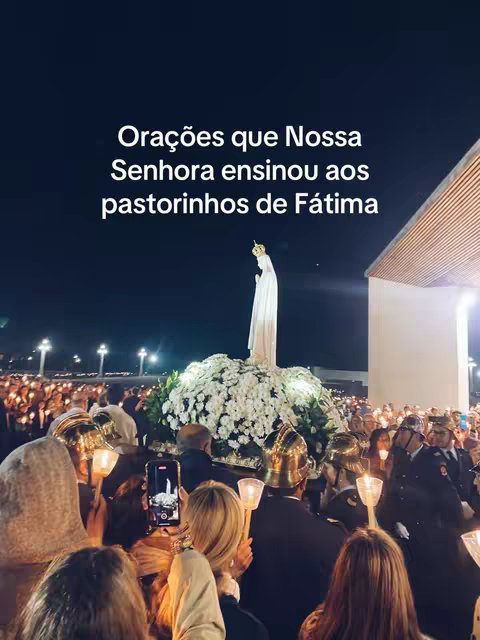 The image shows a nighttime procession with many people holding candles. In the center, a statue of the Virgin Mary is carried atop a vibrant white and green floral arrangement. Text overlays at the top read 'Orações que Nossa Senhora ensinou aos pastorinhos de Fátima' in white text. The scene is warmly lit and appears to be a religious event.