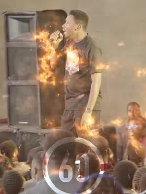 A man with short hair is speaking into a microphone, with flames emanating from his body. The background includes large speakers and a crowd of people with their backs to the camera.  A large timer-like graphic dominates the lower portion of the frame.