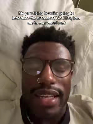 A close-up shot of a Black man wearing glasses, with a neutral expression, resting in a bed covered with a white sheet. The background is a blurred white.