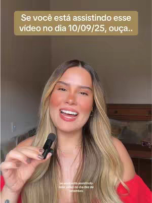 A woman with long blonde hair is holding a microphone and smiling at the camera. She is wearing a red top and a necklace. Behind her there is a wooden cupboard and cream colored wall. Above her is a text overlay reading in Portuguese: 'Se você está assistindo esse vídeo no dia 10/09/25, ouça..'