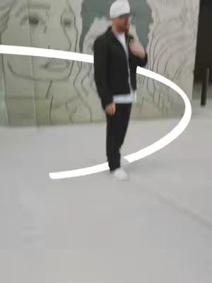 A man wearing a white baseball cap, black jacket, and black pants stands in front of a blurry, patterned background. A white, curved line frames his body. The image has a modern, slightly abstract feel.