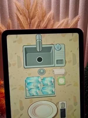 The cover image displays an iPad screen showing a cartoon kitchen scene. The iPad is framed by a blurred background of pink, white, and orange, and some decorative foliage rises above the iPad. The kitchen scene features a sink, dish soap, a stack of blue cleaning supplies and an empty plate. The scene appears to be from a game.