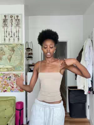 A woman stands in a room, the focal point. She wears a light-colored spaghetti-strap top and white pants. She has dark hair, standing in a fluffy afro. In the background, a map is on the wall, and other decorations give the room a homey feeling.