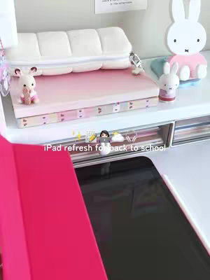 A styled desk setup featuring an iPad with a pink cover and case. Various aesthetic desk accessories are arranged around the iPad, with a light color scheme.