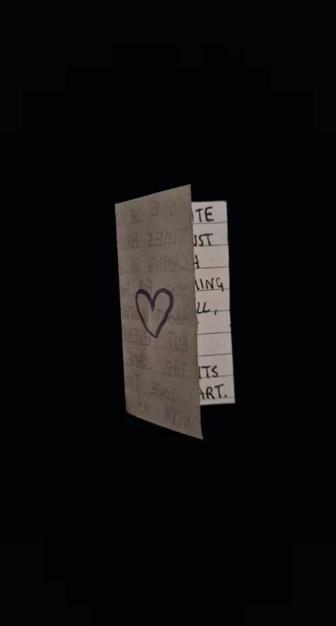 A black background with a small, folded card in the center. The card is beige and has handwriting on it, along with a purple heart drawn with a marker. The handwriting is partially visible and includes the words 'LOVE THE', but it appears to be a loving message to someone.
