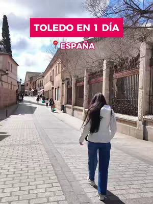 The cover image shows a woman walking away from the camera on a cobblestone street in Toledo, Spain. Overlaid at the top is a red rectangle with white text that reads "TOLEDO EN 1 DÍA", beside a location pin icon and "ESPAÑA". The street is flanked by buildings on either side with a few people walking in the background.