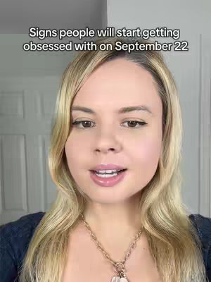 A medium shot presents a fair-skinned woman with long, light-blonde hair. The woman is centered in the image, facing the camera with a slight smile. The background is a white wall and a closed white door. She wears a dark blue, long-sleeved shirt with a gold-colored necklace. Text is overlaid at the top of the image in black font.