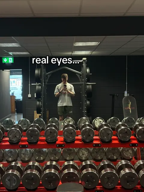 A man is taking a selfie in a gym, reflected in a mirror. He is standing in front of a rack of dumbbells. The text overlay says 'real eyes...' The gym is well lit and has a dark colour.