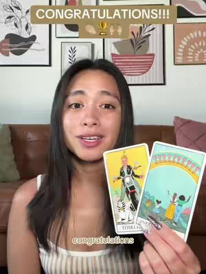 A woman is holding two tarot cards. The background consists of framed artwork and a brown couch. In the upper right corner, there is text overlaid in a beige box with the word 'CONGRATULATIONS!!!'.