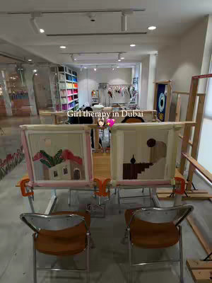 The image is a wide shot of a bright, well-lit studio setup. In the foreground, two framed needlepoint projects are displayed on a table with two metal-framed chairs. Behind the table, there's a workspace with materials on display with a shelf filled with different color threads. The background has a visible rack of clothes, implying a creative or design-focused environment. Overall, the image suggests a crafting or artistic activity.
