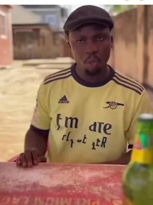 A man in a yellow Arsenal jersey and a cap sits at a table, looking directly at the camera with a slightly quizzical expression. He appears to be in an outdoor setting, with a partially visible bottle in the foreground. The background is blurred, suggesting a street or residential area.