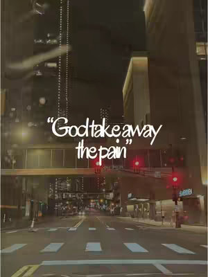 A dusk city street scene. The foreground shows a crosswalk, leading toward multiple buildings illuminated from within. The sky is dark, and the streetlights and building lights are on. At the crossing is a bridge. The text overlay at the top of the image says, “God take away the pain”.