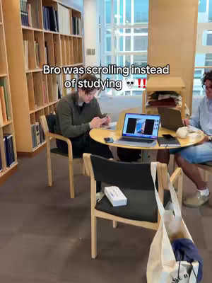 A photo taken in a library setting, showing two figures sitting around a small, round table. There are bookshelves filled with books in the background. One person is using their phone, and the other is using a laptop. The table has a laptop on it.