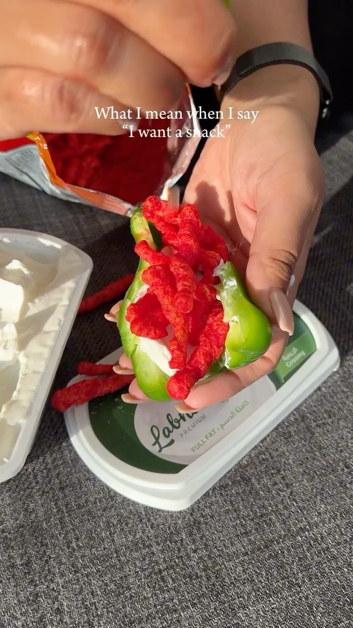 The image shows two hands holding a green bell pepper, which has been sliced open and filled with a white creamy substance and red, crunchy snacks, possibly Flamin' Hot Cheetos. Additional Cheetos are scattered around, and a container of what appears to be a spread or dip sits nearby. The background is out of focus, suggesting a close-up shot. Text overlayed in the image are words that says "What I mean when I say 'I want a snack'"