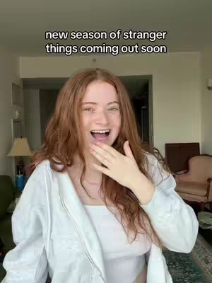 A young woman with long red hair is captured in a selfie-style shot. She is wearing a white top and a matching zip-up jacket. Her mouth is open in a wide smile, and her hand is raised near her mouth in a gesture of surprise. The background shows a room with a window, suggesting a casual, home setting.