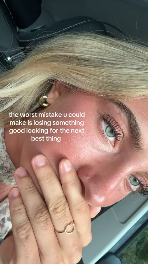 Close-up shot of a blonde woman's face, with a hand covering her mouth. She has green eyes and is wearing a gold ring and earring. Text overlayed reads: 'the worst mistake u could make is losing something good looking for the next best thing'. The background shows part of a car.