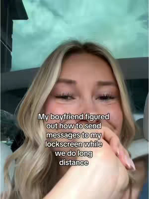 A selfie-style shot from the chest up features a young woman with long, wavy blonde hair. Her expression is one of gentle happiness, with a slight smile and eyes looking down, possibly in amusement or shyness. She is holding her hand up to her mouth, with light pink nail polish visible, perhaps hiding a giggle. The background is blurred, suggesting the inside of a vehicle with a glimpse of a cloudy sky visible through the window. The lighting is soft and natural, highlighting her features. Text is overlaid on the image.