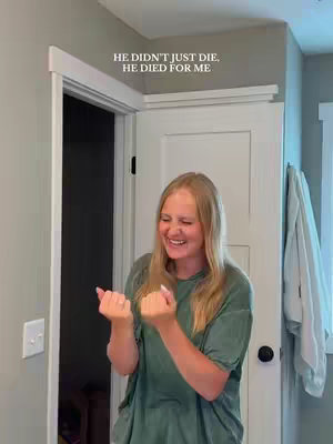 A woman with blonde hair is smiling and laughing near a doorway in a bathroom. The video seems to be filmed in a home environment. The woman is wearing a green t-shirt and is gesturing enthusiastically with her fists. Above the doorway is white text that reads: HE DIDN'T JUST DIE, HE DIED FOR ME.