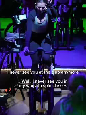 A woman in workout gear is riding a stationary bike in a cycling studio at a worship workout. The image is well-lit with a dynamic composition, with vibrant lighting and a focus on the woman.