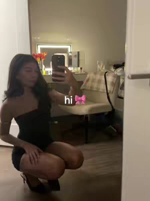 The cover image shows a young woman posing for a mirror selfie. She appears to be in a well-lit room, possibly a hotel room or a similar setting. The woman is wearing a black, strapless mini dress and high heels, suggesting a night out or special event. She is holding a phone in her hand, likely to take the picture, and a pink bow drawn on the image, and the text 'hi'. The background shows a blurred view of the room, including a bed, a mirror, and some decorative elements.