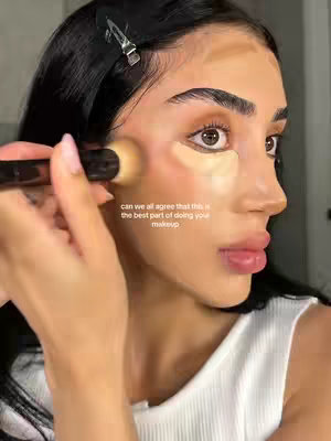 A close-up shot features a person applying makeup. The person, with dark hair pulled back, is using a makeup brush to blend makeup on their cheek, with visible contouring and highlighting. The background is blurred, keeping a focus on the makeup application. The overlay says 'can we all agree that this is the best part of doing your makeup'.