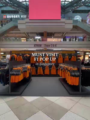 An indoor shot of an F1 merchandise store in a mall. The foreground features displays of orange and black jackets and other apparel. The background includes escalators, a large pink digital display, and store interiors. The text overlay promotes the store.