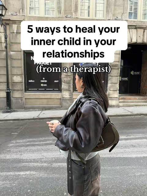 A woman stands in front of a building, her side profile visible. She holds a shoulder bag and wears a brown jacket. A text overlay in a speech bubble shape reads: "5 ways to heal your inner child in your relationships (from a therapist)". The background is a city street with the building, a street lamp, and storefront signage.