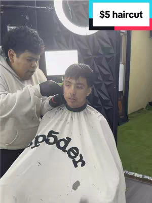 A person is getting a haircut by a barber, shot from a point-of-view angle. The barber is wearing a white sweatshirt, and the person getting the haircut is wearing a white cape.