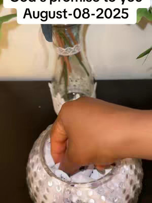 The image shows a hand reaching into a glass bowl filled with small white slips of paper. The bowl is filled with reflective surfaces and rests on a dark surface. Beside the bowl is a vase with a delicate arrangement of flowers. The background has elements from the vase and a dark countertop.