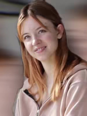 A young woman with long auburn hair smiles gently while facing the camera. She is wearing a light pink zip-up hoodie. The background is softly blurred.
