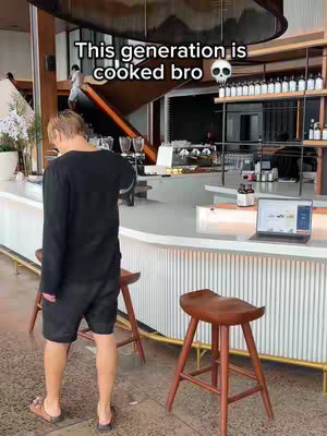 A man stands with his back to the camera in a modern cafe setting. He faces a white counter with stools, a laptop, and shelves with products. The scene is mostly neutral tones with wood accents and good lighting.