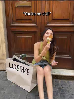 A woman sits on stone steps in front of a wooden door holding an ice cream cone, a LOEWE shopping bag is to her left. Text overlayed on the image reads "You're so chill".
