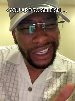 The image is a close-up shot of a Black man with a concerned expression. He is wearing a gray baseball cap and a white patterned short-sleeved shirt. The text overlay says, "YOU ARE SO SELFISH...". He is looking off to the side with his mouth slightly open.